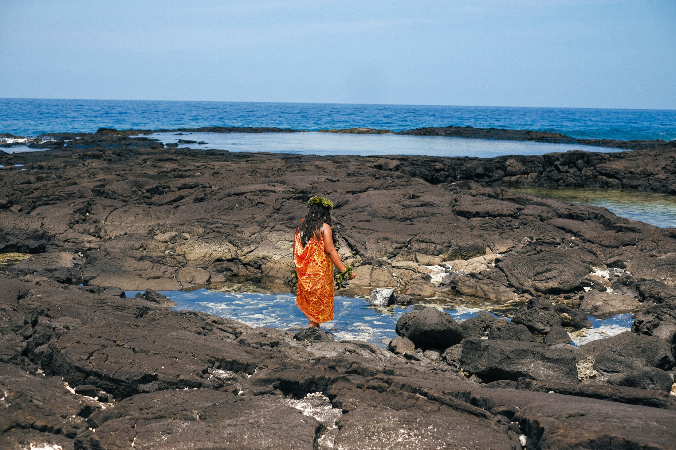 A woman in a kīhei and haku lei crossing the black lava shore toward the sea